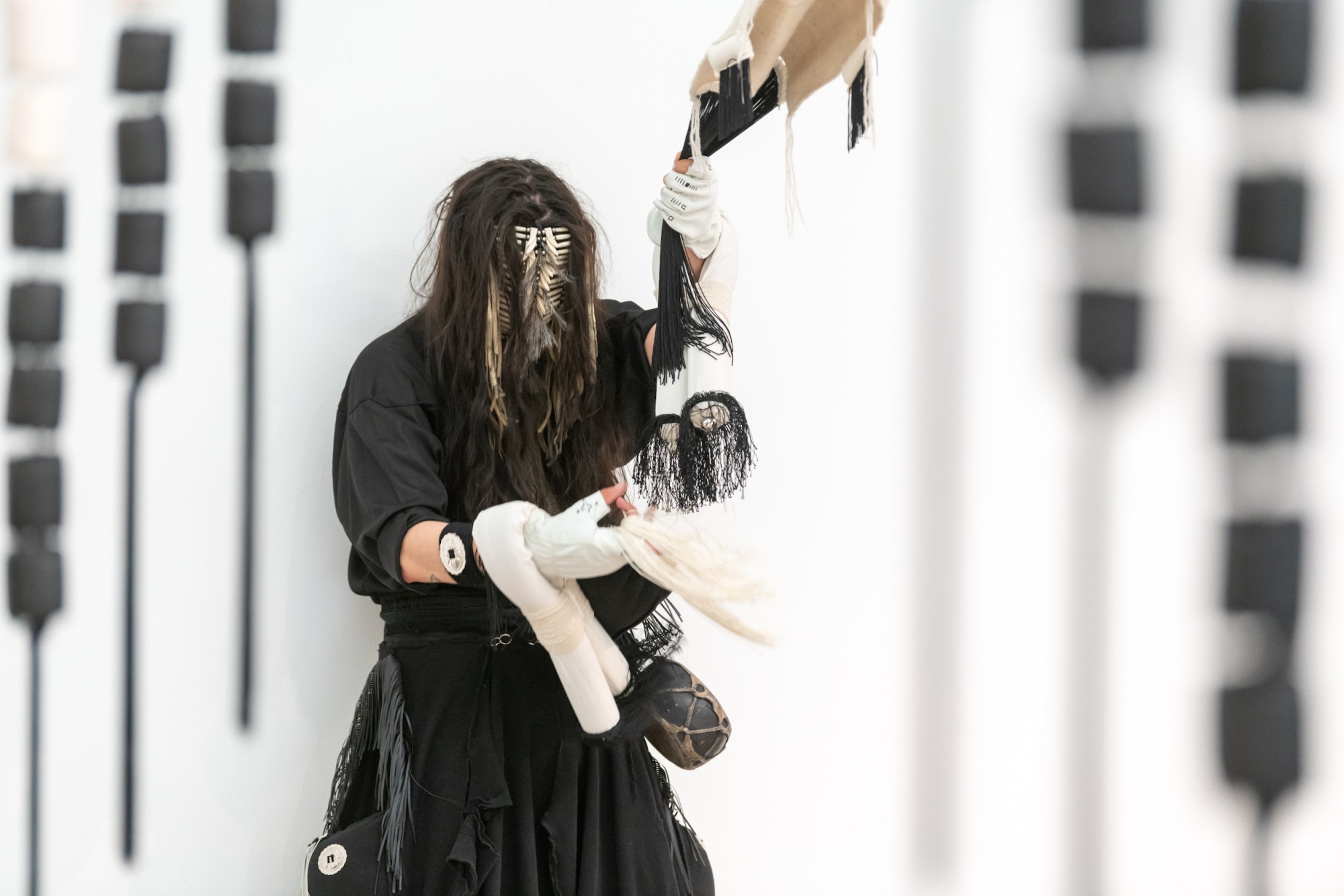 Artist Eric-Paul Riege dancing with a white mask, loose black clothes, and several large textile-based ornaments in his hands.