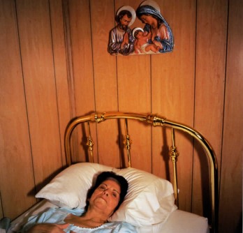 A sick woman lying in a small bed with a gold bedframe in a wooden room with Joseph, Mary, and Jesus hung on the wall at her head