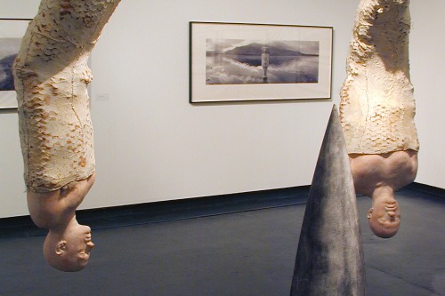 In a gallery two photographs hang on the wall. In front of those, two sculptures of people hang upside down in what looks like cocoons above clumps of fur. In front of them, a large spike-like object sits on the floor