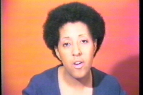 A black woman wearing a blue shirt in front of a yellow, red, and pink ombre background is mid sentence, with a slightly old-television-like filter