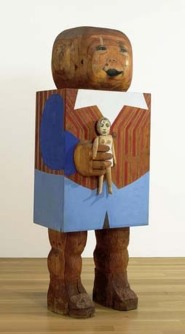 A large wooden doll with a rectangular body holds a smaller, more humanoid doll