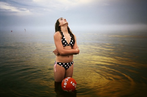 A woman with brown hair in a polka-dot bikini stands in a lake with a red volleyball looking up to the sky
