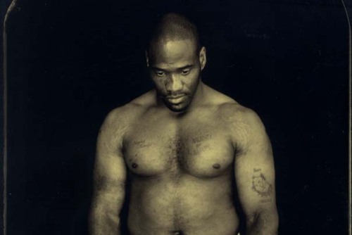 A black and white photo of a shirtless black man in jeans looks down with his hands by his side