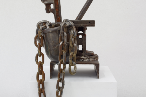 A rusty metal sculpture of wrenches, chains, chisels, and other metal items sits on a white pedestal in a white room
