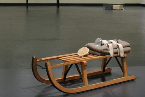 A sled with a few objects on it sits in a gallery in front of a glass box on a table and an installation on the ground