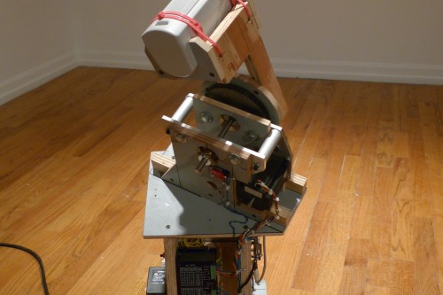 A mechanical sculpture made from plywood, bolts, zip ties, and general machinery sits in a white room with a wood floor plugged into a wall off picture 