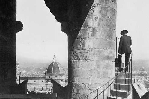 A black and white photo of a man going up a spiral staircase on a pillar on top of a building