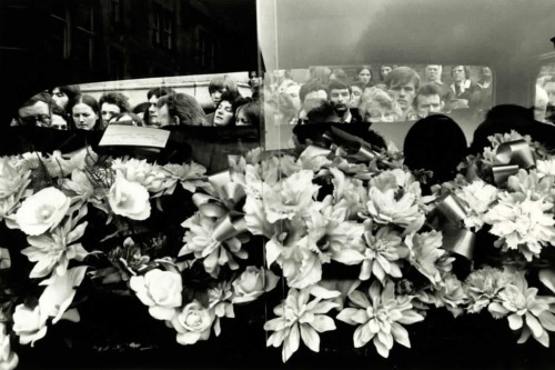 A black and white photograph of people crowded around a car containing white flowers