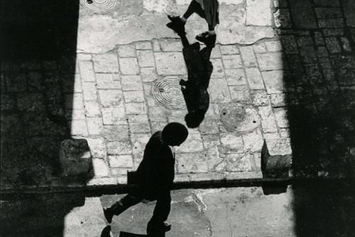 A black and white photograph taken from above shows a man and a woman walking on the road