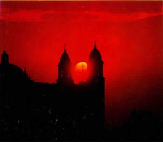 The silhouette of an old building against a red sky with the sun setting between two of the towers