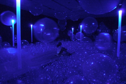 A person sits on a thin platform in a blue room full of large plastic balls 
