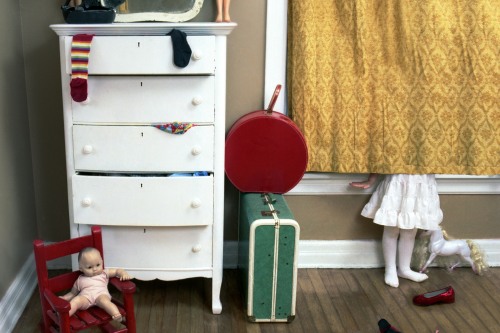 A girl in a white dress stands behind a gold curtain next to a suitcase and white cabinet, with toys about