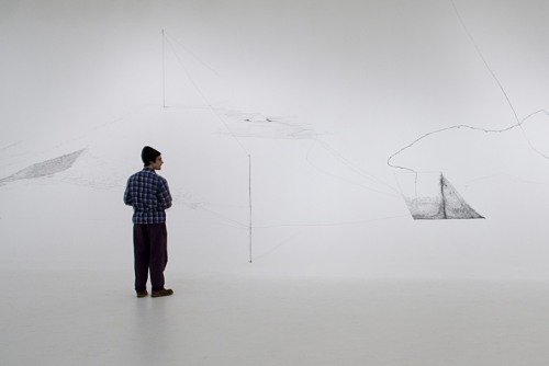 A man in dark clothing stands in a spare white room with drawings on the walls