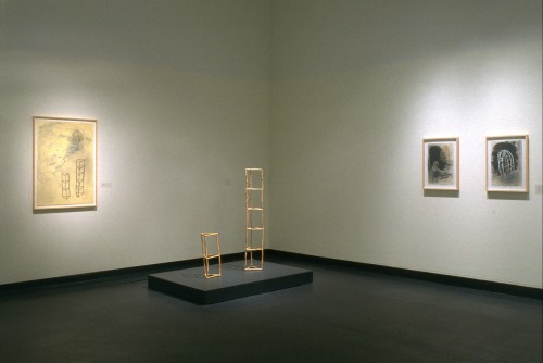 A painting of two scaffold like objects sits on a gallery wall, and the objects in the painting are installed in front of the painting. There are two smaller paintings to the left 