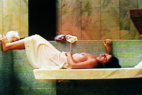 A naked woman wearing a towel lies on a platform in a large bath room with green tiles and marble walls