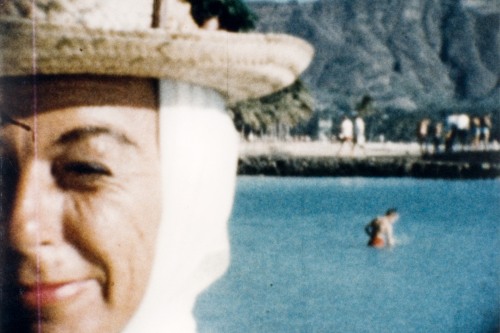 A woman squints in front of the camera and behind her a man wades in the water in front of a mountain