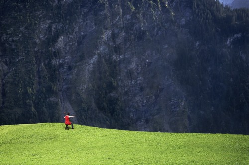 Person wearing red plays the cello on a green hill facing a mountainside