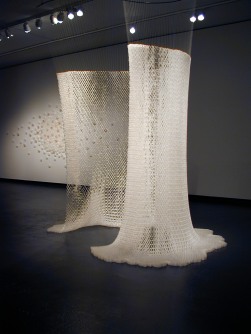 Installation view; a sculpture of two large white fabrics hanging from the ceiling with a bright light cast upon them