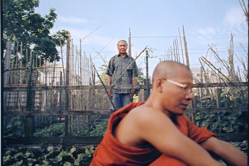 Photograph of two men; one monk in the foreground out of focus and a farmer in the background focused and looking at the camera