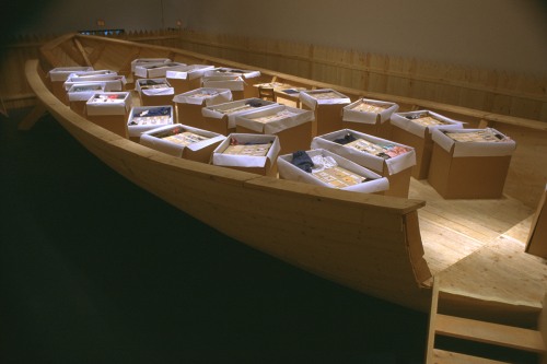 Installation view of a large wooden boat containing cardboard boxes with various items