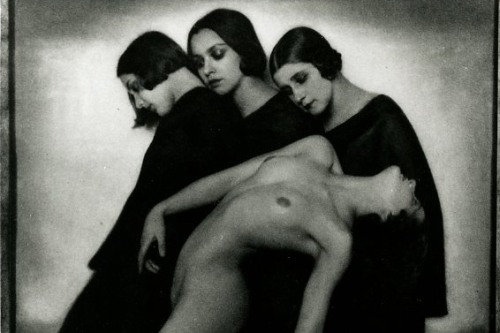 Photograph of three women in black gowns, and a nude woman standing in front of them leaning backwards