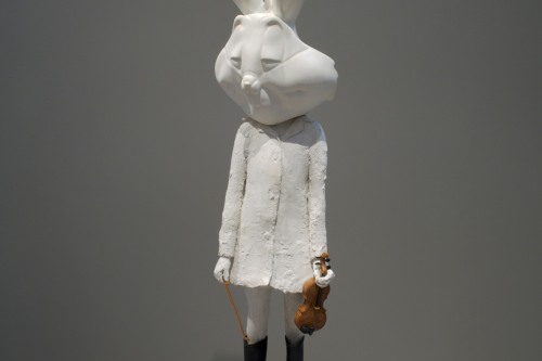 Installation view of a sculpture of a human figure with a rabbit head, holding a violin, standing on a green step stool