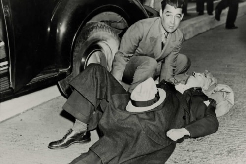 Black and white photograph of two men; one is laying on the street and the other is leaning over him, looking at the camera