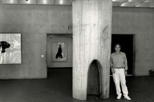 A black and white photo shows a man leaning on a thick hollow pillar with pictures hanging on concrete walls behind him