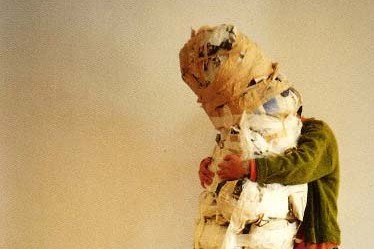 A man in a green jacket and black pants hugs a large wrapped object in a white room with wood floors
