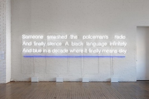 LED light text on a white wall reads "Someone smashed a policeman's radio And finally silence A black language infinitely  And blue in a decade where it finally meets sky"