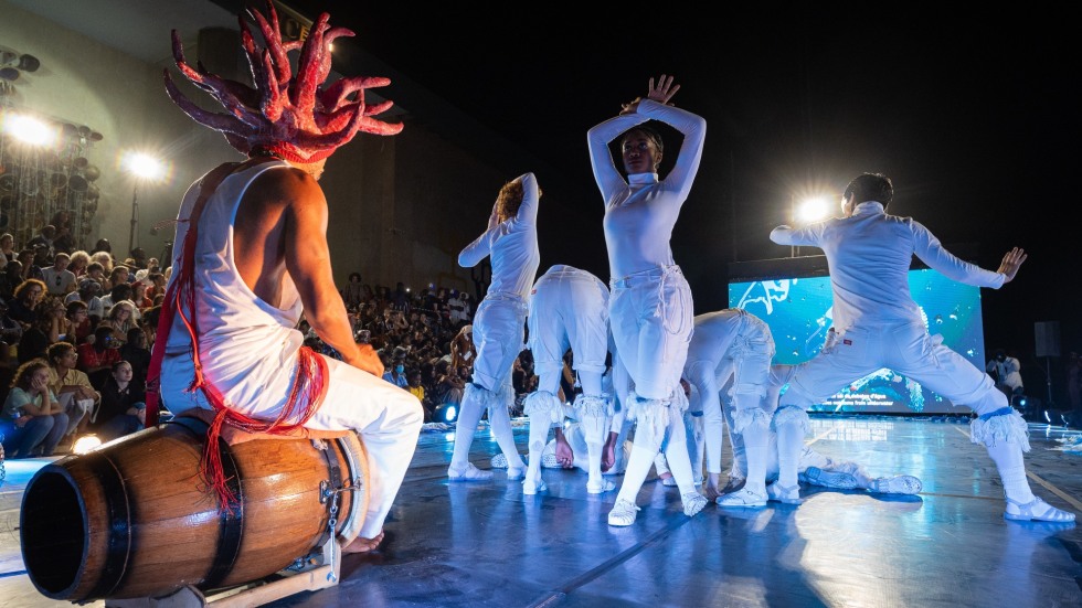 A group of dancers dressed in all white striking various poses in center stage. To their left is a figure sitting on a drum with a red coral-like headpiece.
