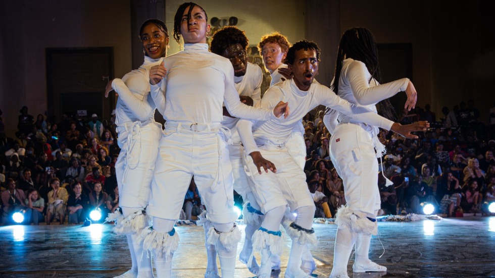 A group of 6 dancers dressed in white with their arms bent in various poses and a wide range of facial expressions.