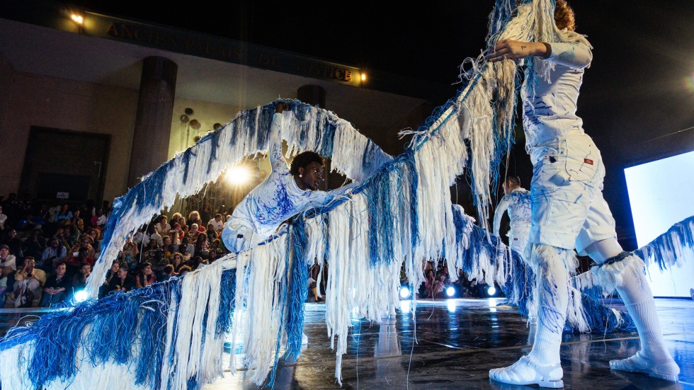 Two dancers dressed in white lifting flexible poles draped in white and blue tassels.